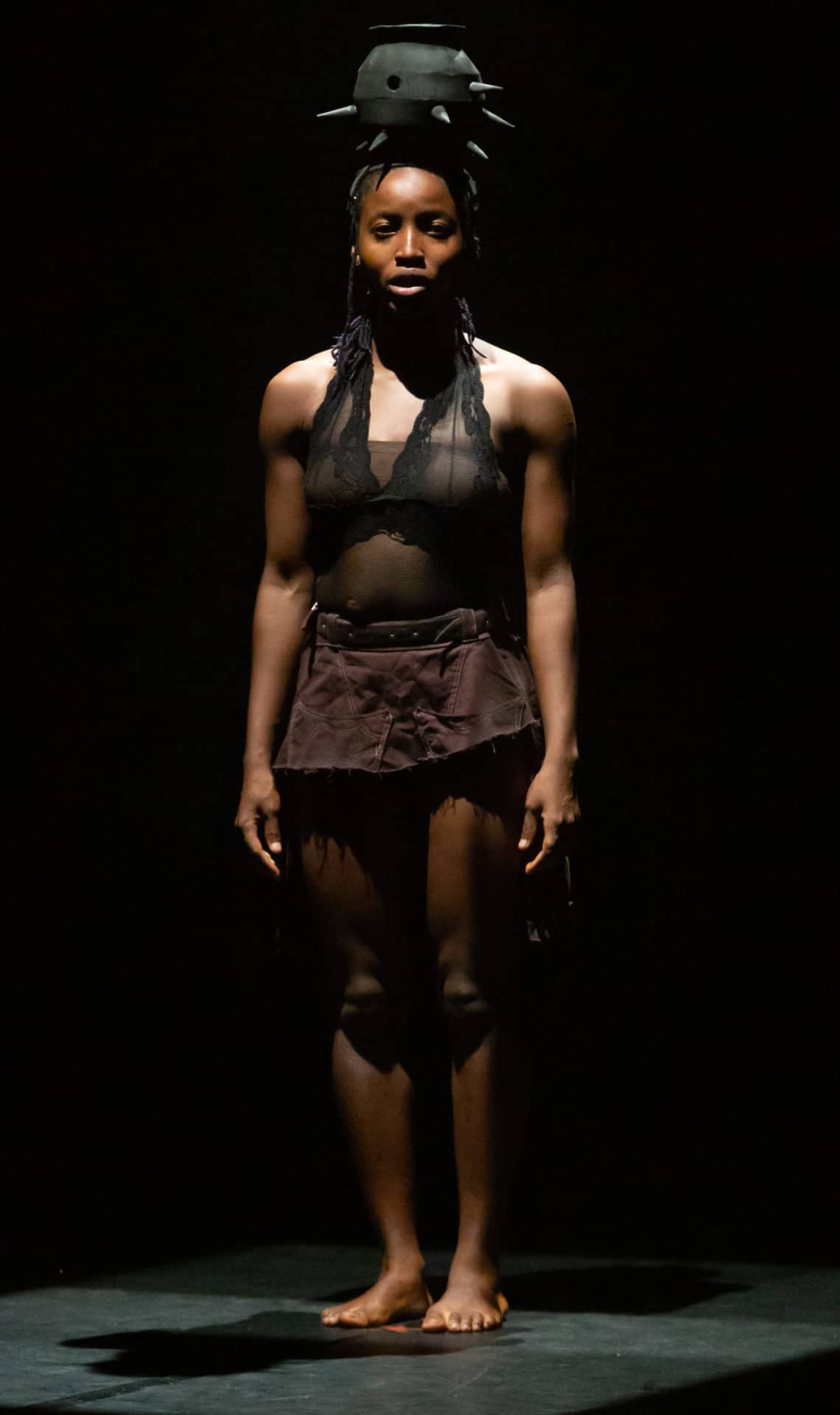 A performer in QDance Company stands tall, balancing a spiked iron cauldron on her head, in the light on a darkened stage at the Centre Pompidou in Paris, France. Press image courtesy of PS21