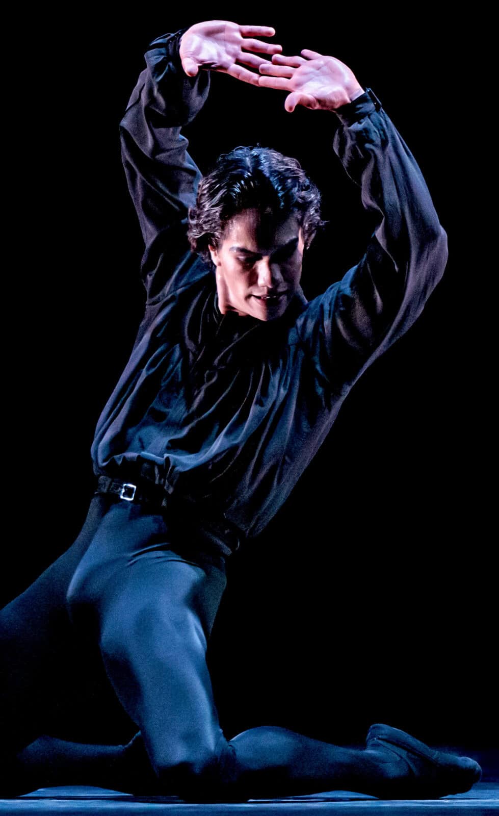 A dance artist with San Francisco Ballet lifts his arms in black cloth in the dark. Press photo courtesy of Jacob's Pillow