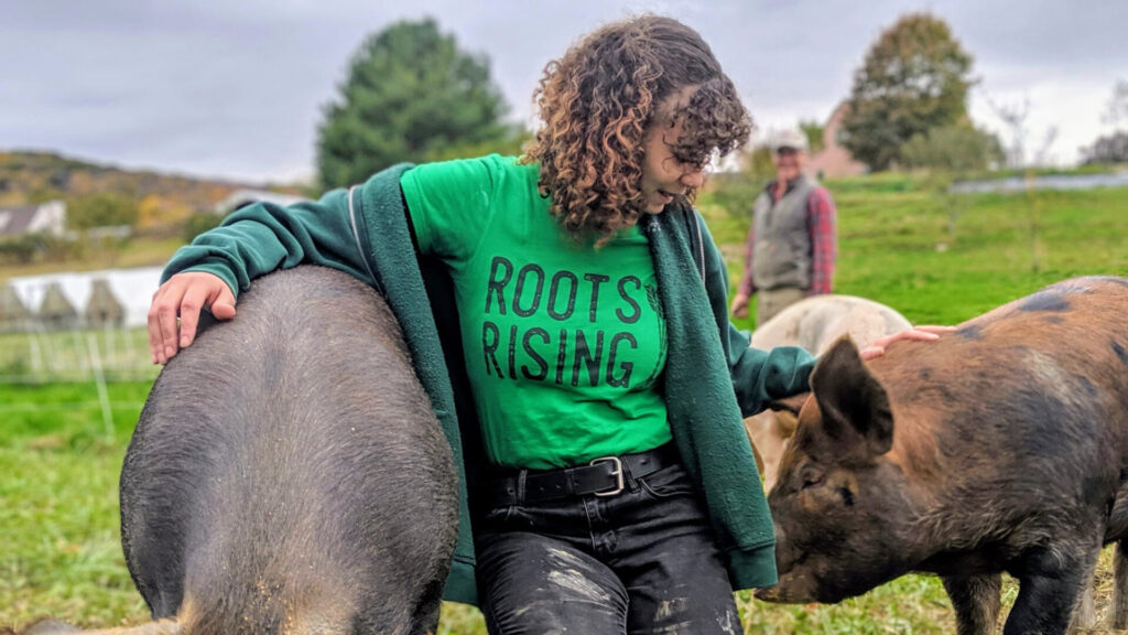 A teen member of the Roots Rising farm crew hangs out with contented young pigs. Press photo courtesy of Roots Rising.