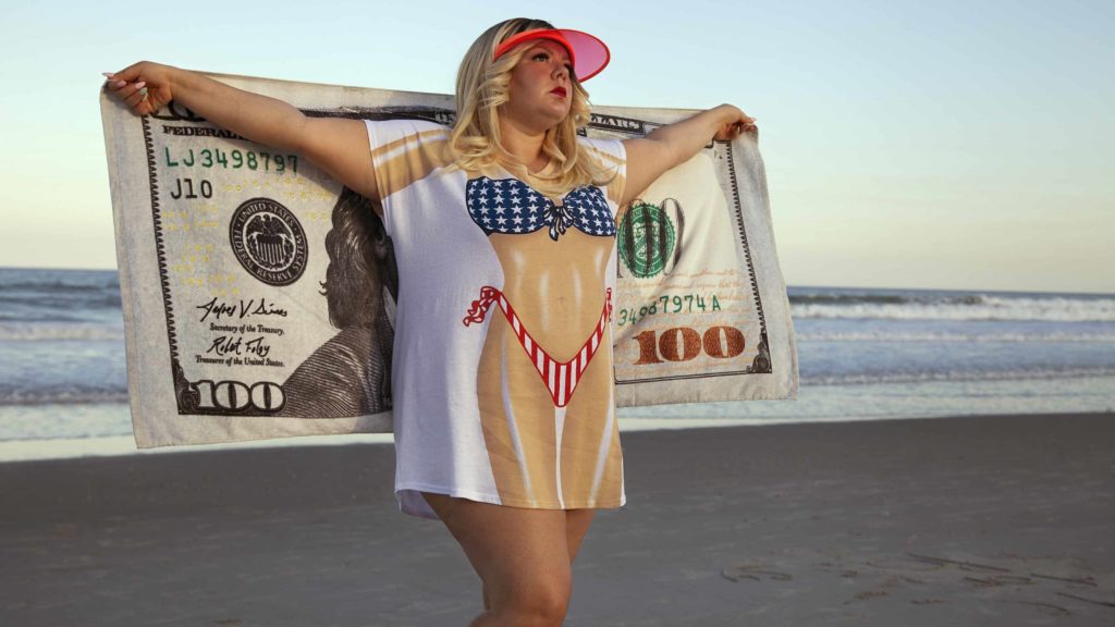 Genevieve Gaignard poses on the beach with a towel printed as a $100 bill and a t-shirt painted with a svelt body, in her self-portrait 'Counter Fit,' courtesy of the artist and MCLA Gallery 51.