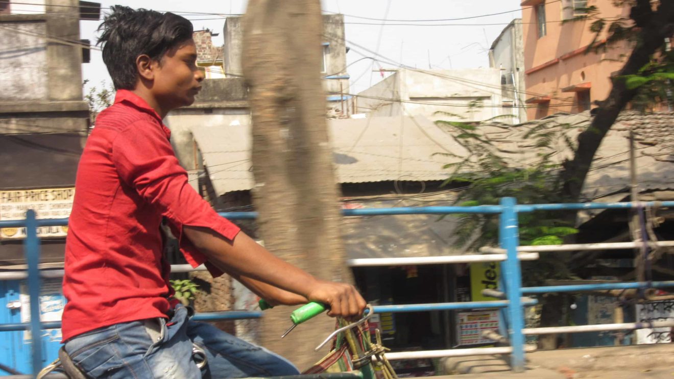 A man in a red shirt and jeans rides a bicycle in Kolkata, India.