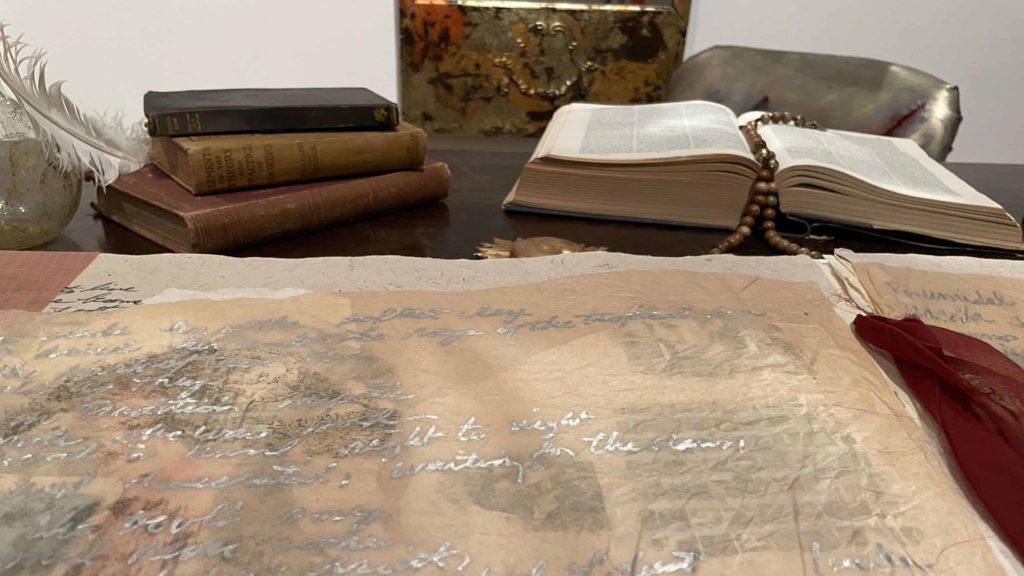Long scrolls of paper unroll on a desktop in a closeup from Amalia Mesa-Bains' installation, The Library of Sor Juana Inez de la Cruz at WCMA