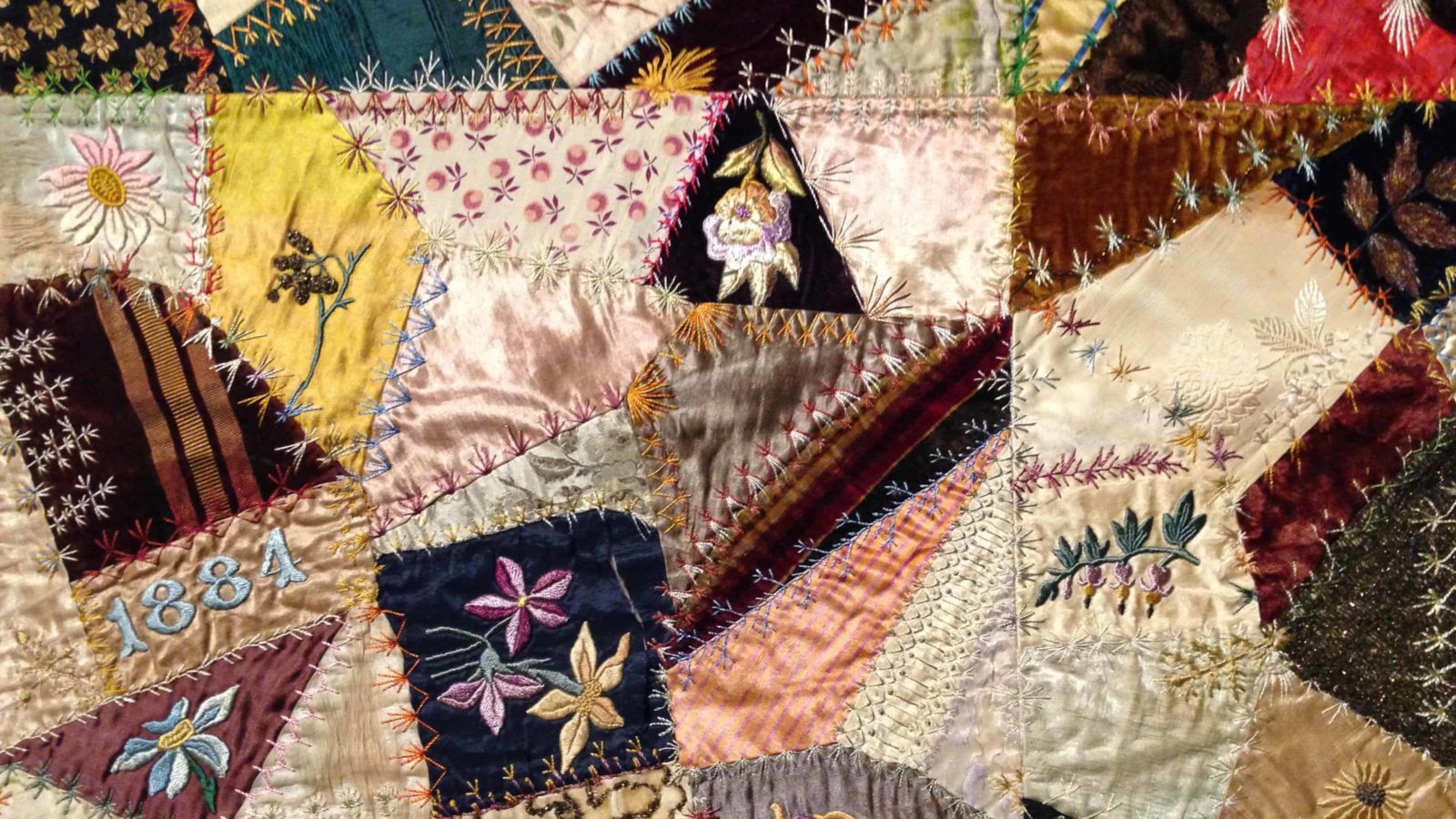 A close-up of a crazy quilt at Bennignton Museum shows bright, scraps of cloth in varied colors and textures, with patters of stitching and embroidered flowers.