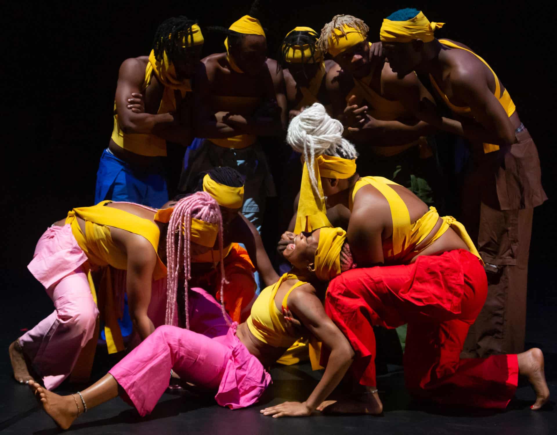 Performers in QDance Company gather in the light, in bright costumes on a darkened stage at the Centre Pompidou in Paris, France. Press image courtesy of PS21