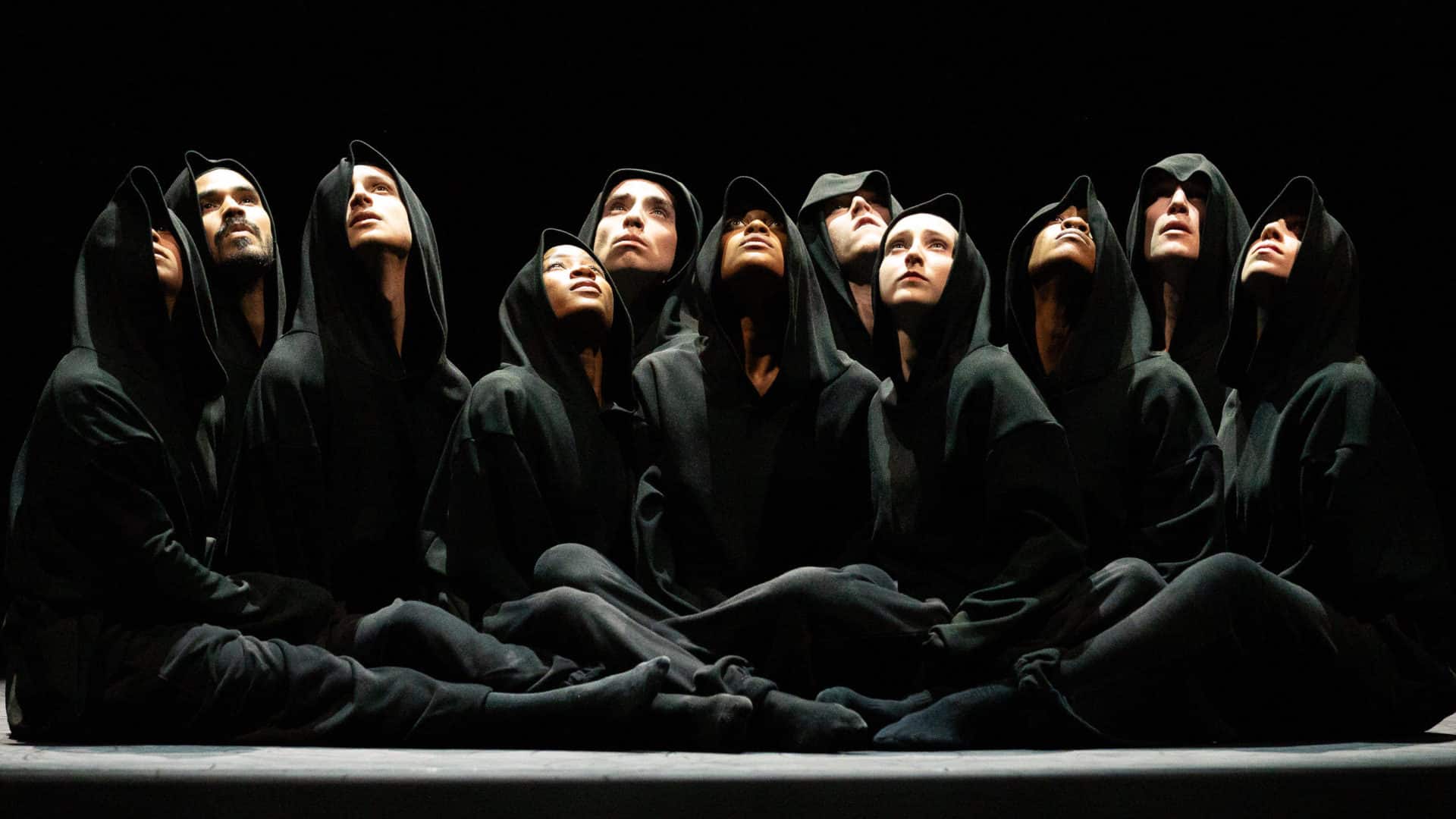 Hubbard Street Dance Chicago dancers sit close together in dark hoodies, looking up into the light in Azshure Barton's Busk at Jacob's Pillow. Press photo courtesy of Jacob's Pillow