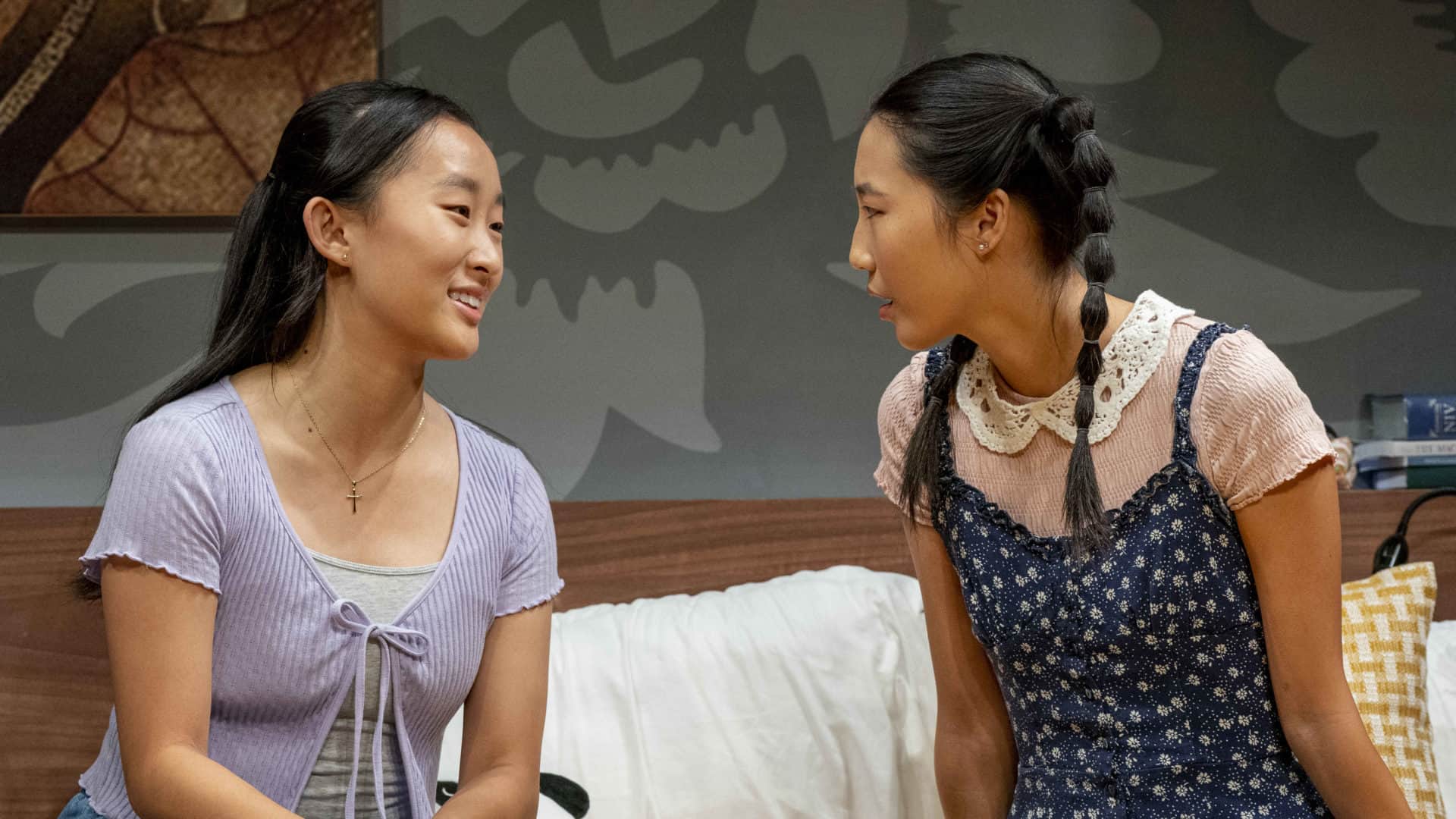 Ji-young Yoo and Shirley Chen siton a hotel bed, smiling, in Man of God by Anna Ouyang Moench. Press photo courtesy of Williamstown Theatre Festival
