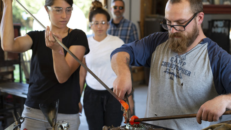 Artists in a glassblowing workshop turn glowing molten glass on long rods at Salem Art Works. Press photo courtesy of SAW