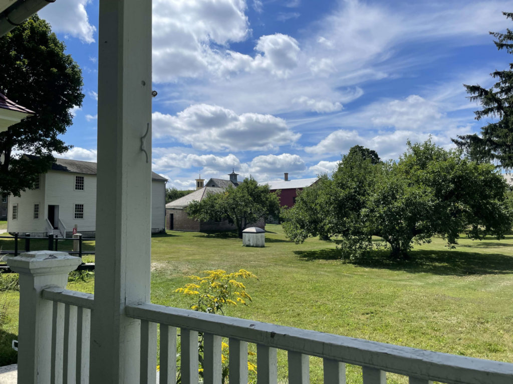 From the porch of the ovvice building, a guest on a rocking chair can look across Hancock Shaker Village.