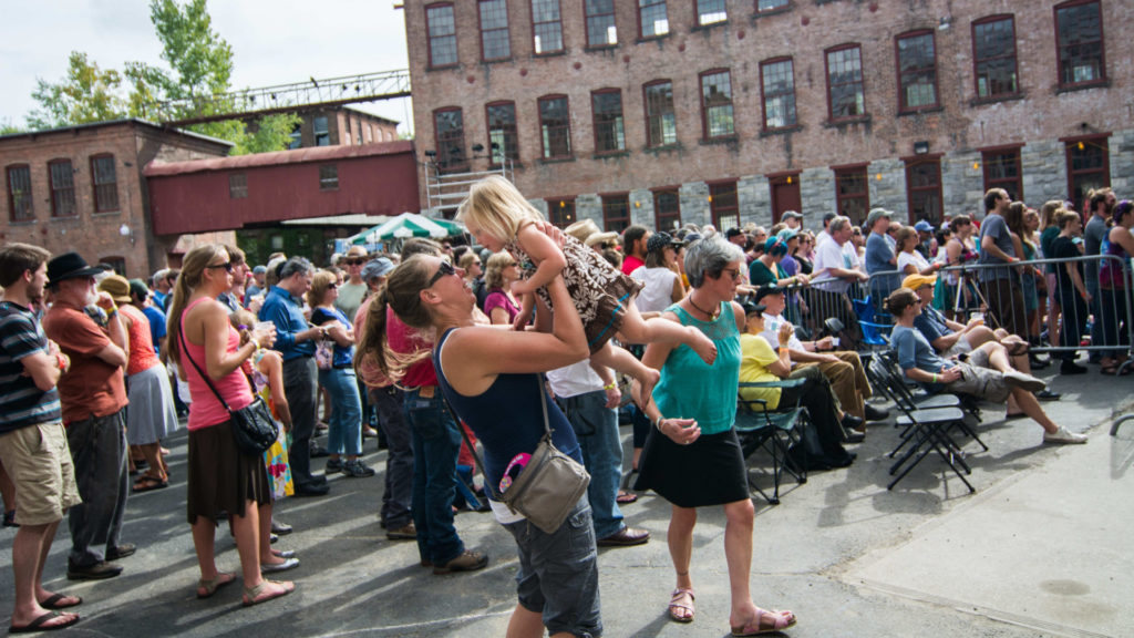 A woman lifts a child in a courtyard full of people listening to live music at FreshGrass. Press photo courtesy of Mass MoCA