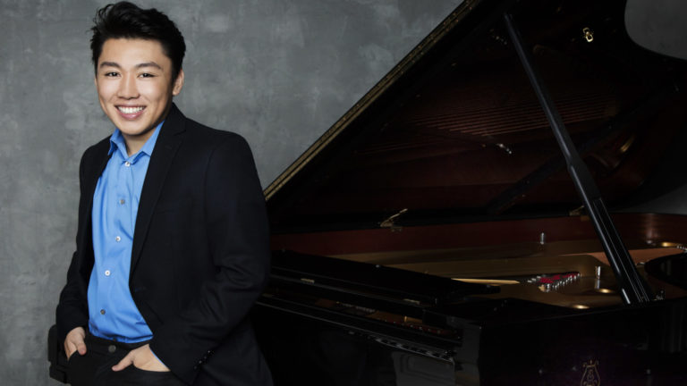 Pianist George Li stands smiling beside a piano. Press photo courtesy of the artist