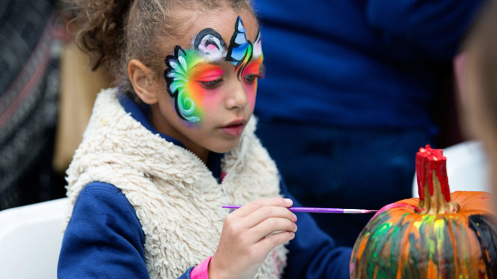 A girl with a vivd rainbow butterfly painted on her brow paints a pumpkin at the Berkshire Botanical Garden's annual Harvest Festival. Press photo courtesy of BBG