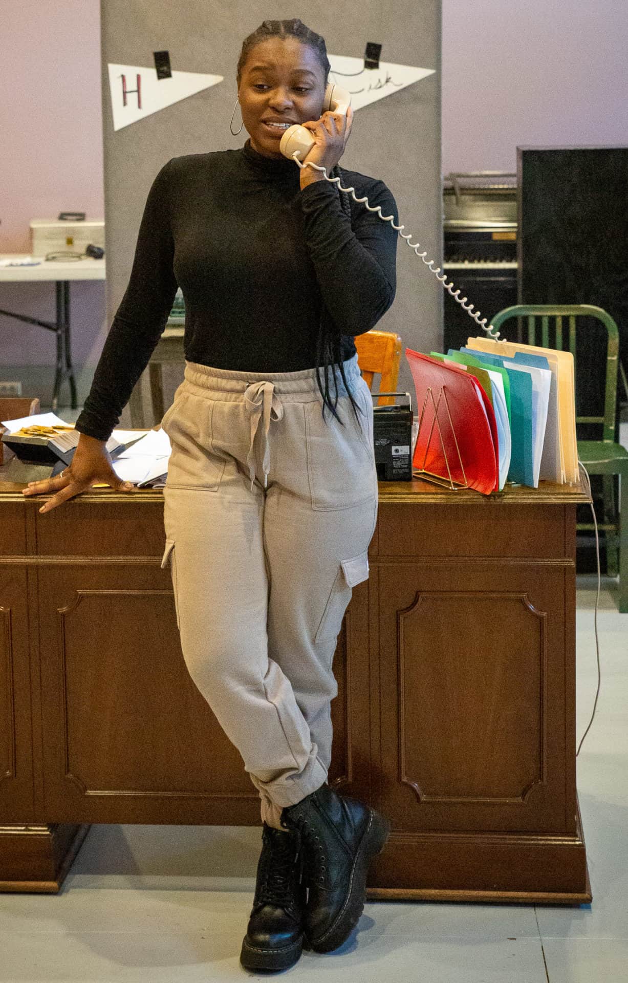 Kyra Davis stands fronting her desk and answering the phone in Cadillac Crew with WAM Theatre. Press photo courtesy of the theater