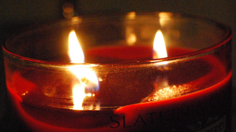 A dark red candle in a glass cup glows with two wicks. Creative Commons ourtesy photo.