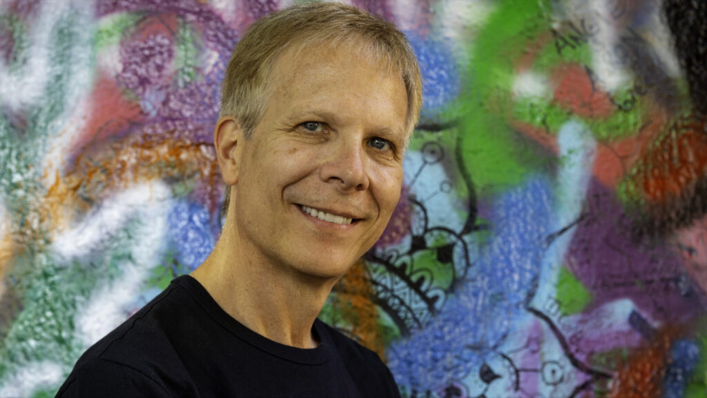 Terezín Music Foundation director Mark Ludwig stands smiling in front of a colorful mural.