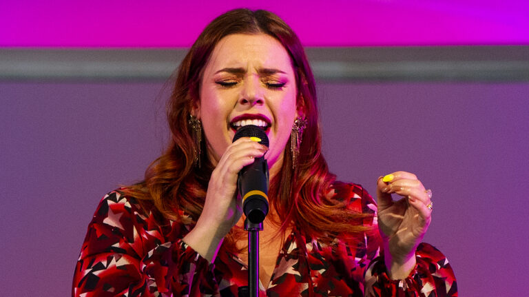 Actor and vocalist Alysha Umphress sings into a microphone at Barrington Stage Company in pink and purple stage lighting. Press photo courtesy of BSC