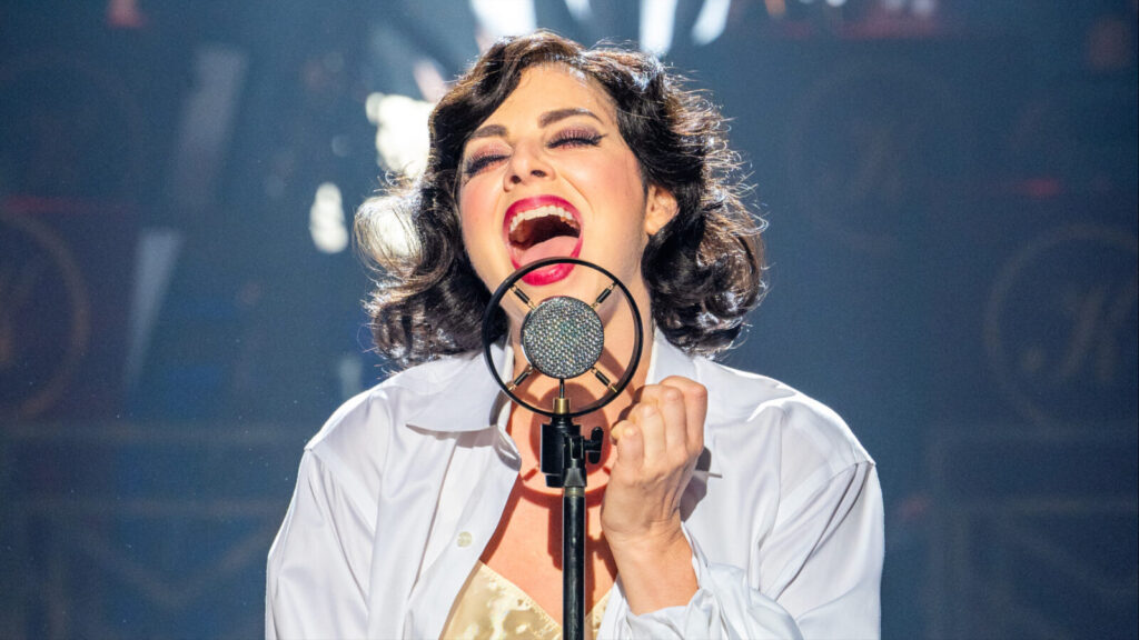 Krysta Rodriguez as Sally Bowles sings into a mic in vivid light in Cabaret at Barrington Stage Company. Press photo courtesy of BSC