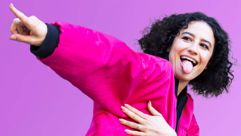 Comedian Ilana Glazer points up left in a vivid pink shirt and sticks out her tongue with a laugh. Press photo courtesy of Mass MoCA.