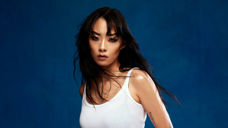 Japanese British pop star Rina Sawayama stands in a white tank top against a blue background wtih her hair down around ehr shoulders. Press photo courtesy of Mass MoCA