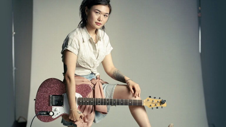 Acclaimed electric guitarlist stands casually holding her guitar. Press photo courtesy of PS21