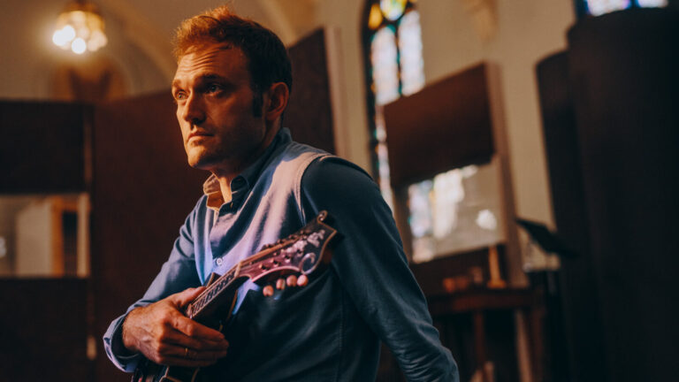 Internationally acclaimed mandolinist Chris Thile holds his instrument in the light from a stained glass window. Press photo courtesy of the artist