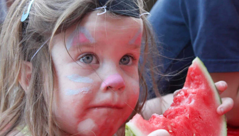 A young visitor with cat face paint samples a slice of watermelon as Third Thursday brings families to North street in Pittsfield.