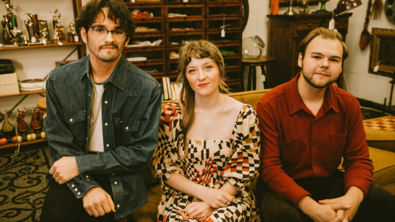 Folk/americana trio The Wildwoods — Noah and Chloe Gose on guitar and violin and Andrew Vaggalis on bass —sit together on a yellow couch in an antique shop. Press photo courtesy of the artists.