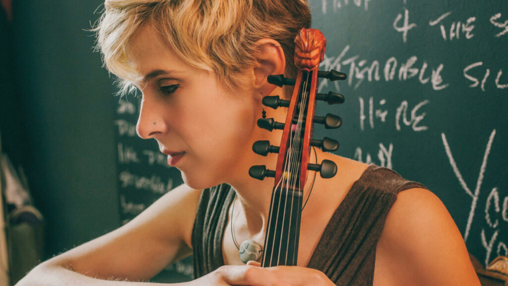 Grammy nominated jazz violinist Sara Caswell sits with her instrument against a green chalkboard. Press photo courtesy of Lulufest and the artist