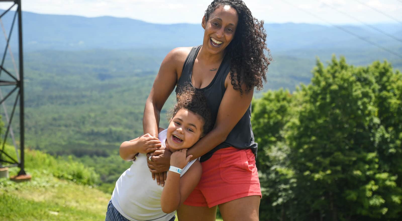 Rachel Hailey, founder of DEI Outdoors, and her daughter, Aria, laugh together on a ridge in the sun, with a wide view over the valley.