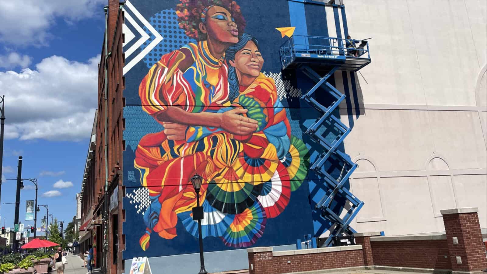 Two women hold each other in a painting a building high, as Sylvia López Chavez' mural Sisterhood brings vivid color to North Street in Pittsfield.