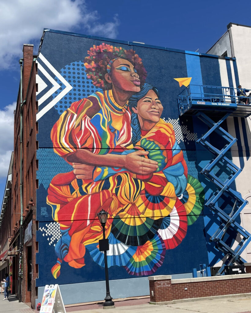 Two women hold each other in a painting a building high, as Sylvia López Chavez' mural Sisterhood brings vivid color to North Street in Pittsfield.