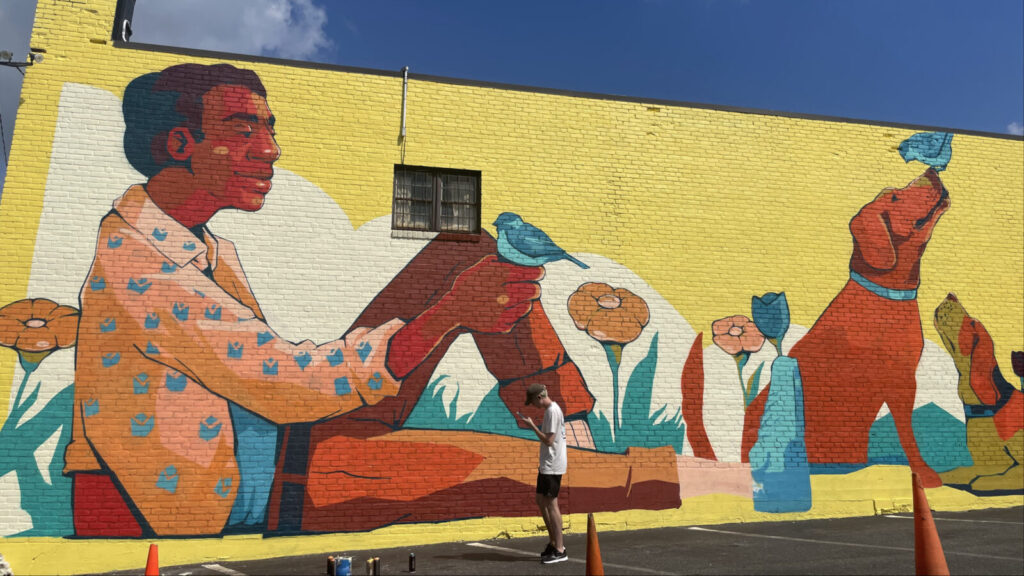 Bluebirds flock around a man and a dog on a sunny yellow background in Qwynto's mural on the side of Carr Hardware on North Street in Pittsfield