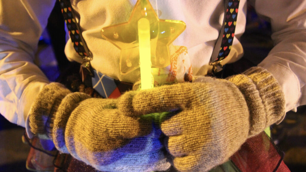 A Halloween trick-or-treater holds a glow stick in gloved hands.