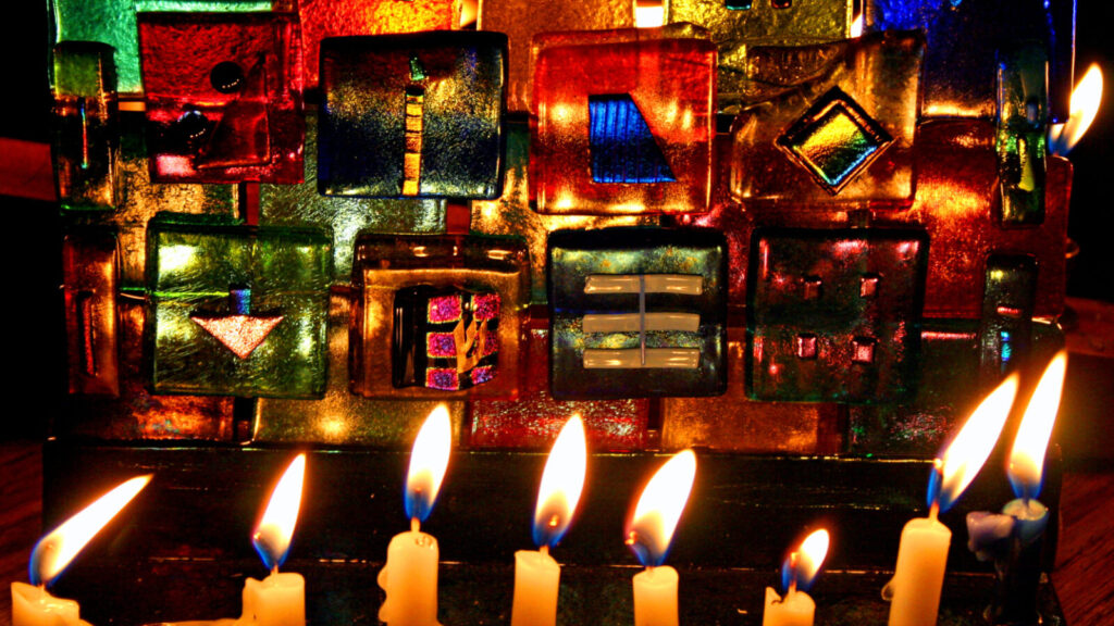 Candles glow in a menorah against a background of bright colored glass. Creative Commons courtesy photo