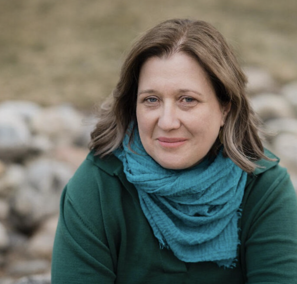 WAM Theatre's Associate Artistic Director, Talya Kingston, sits smiling in a blue scarf with stone and grass behind her. Press image courtesy of WAM