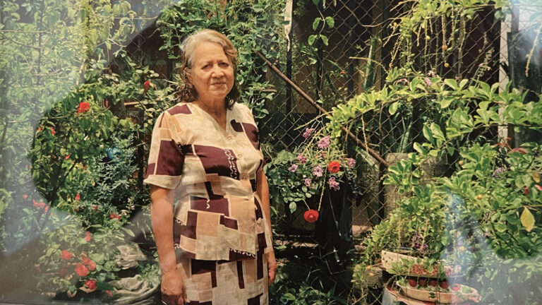A woman stands in her garden on a sunny day, surrounded by flowering bushes, in Lorena Molina's photographs in 'Unfortunately It Was Paradise' at MCLA in North Adams.