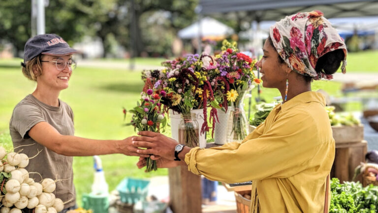 Two women, a farmer and a local neighbor, meet over a table of flowers at the Pittsfield Farmers Market. Press photo courtesy of Roots Rising